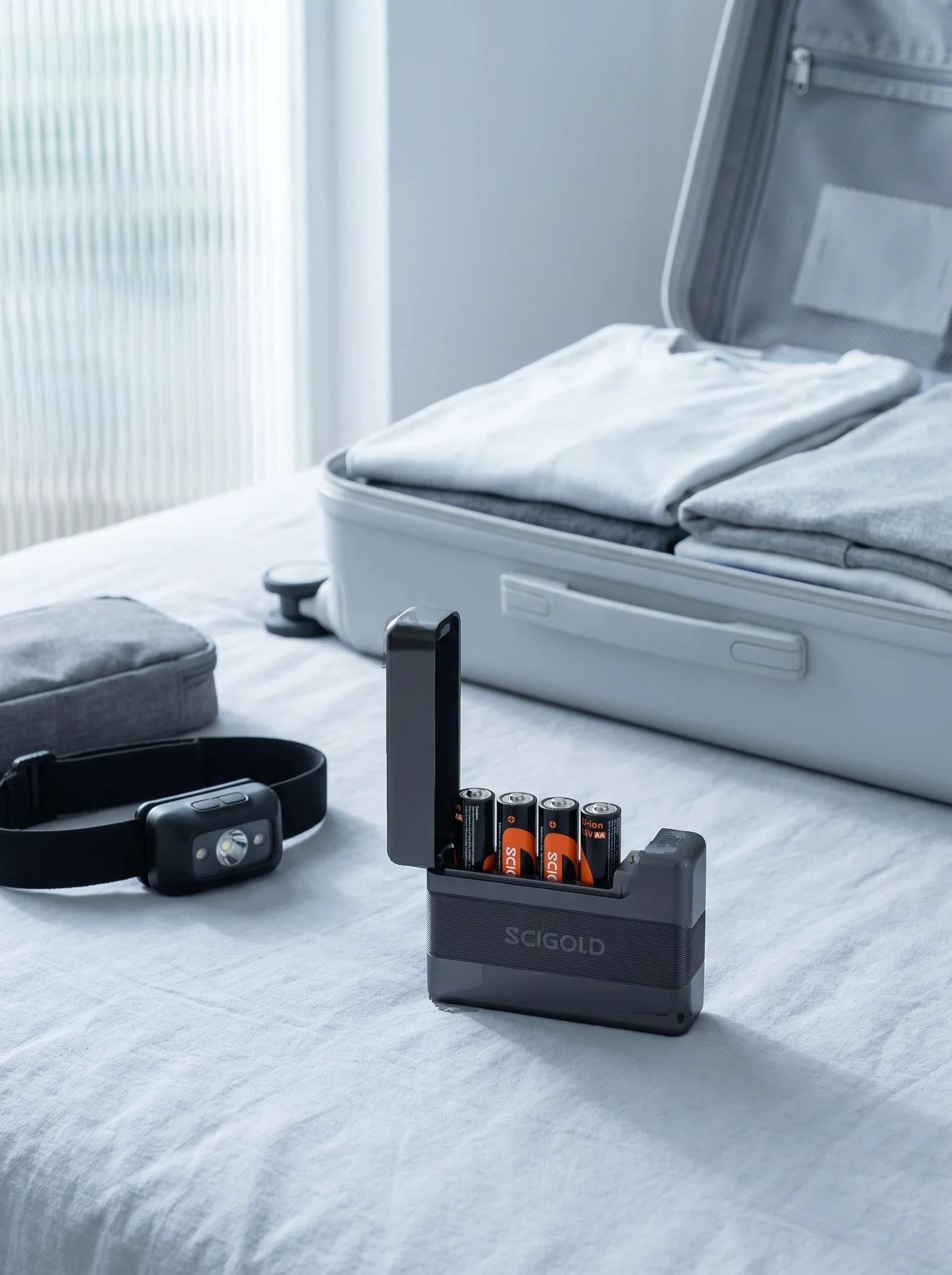 SCIGOLD Pocket Charging Case with four AA cells on a bed beside a headlamp and an open carry-on